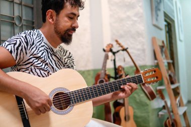 A Latino male musician quietly plays his acoustic guitar. He is dressed in a striped shirt. There are some ukuleles in the back of the room.