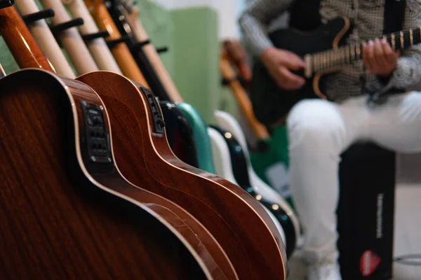 Many electric and acoustic guitars on a stand in the foreground with a musician playing in the background. Body of the guitars. Guitars on Music Store. Musician playing a son in the background with