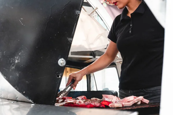 Side view of a grill full of meat and a girl working. High quality photo. Girl in butcher shop grilling meat. Grill with beef and sausages. Traditional roast meat. Unrecognizable girl working in the