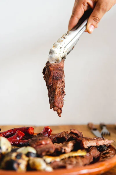 A cut of meat in a tong is about to be placed on a plate. High quality photo. Roasted onion, cuts of meat, and sausages in a dish made of clay. White background. Hand with a pliers