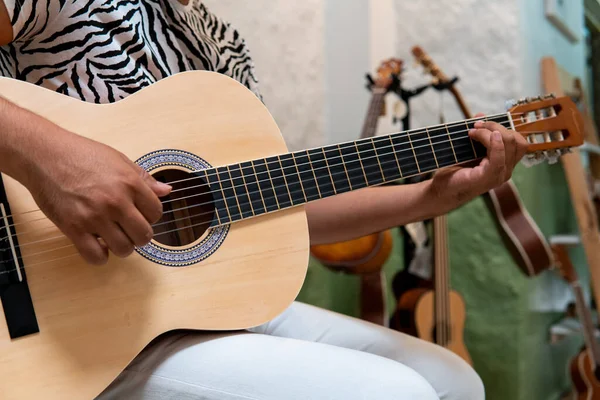 Close-up detail of a brown acoustic guitar and the hands of a musician playing a song. High quality photo. Ukuleles in the background. The musician is wearing a black and white outfit. Green and white