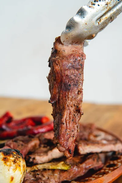 Piece of meat in tongs on a plate full of meat onions and sausages. . Wooden table and white background. Delicious cut of meat. Traditional dishes. Close up to the. Meat.