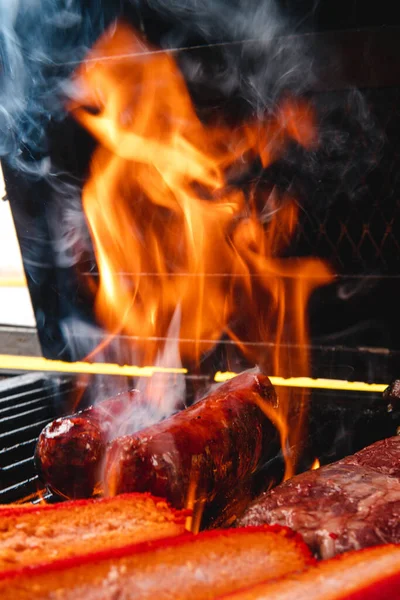 A sausage being grilled with full fire and smoke. There are whole and cut sausages. There is the black grill in the background. Close-up shot of a sausage on fire with the spit.