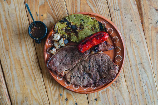 Dish with top sirloin meat cut accompanied by nopales, onions, grilled sausages and sauce on a wooden background. High quality photo. Mexican dishes. Traditional barbecue. Meal from grill.
