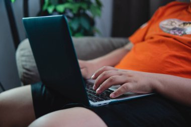 Evdeki koltukta bilgisayarını kullanarak oturan tanınmayan kişi. Kişi turuncu kıyafetler giyiyor. Arka planda bitkiler ve kahverengi bir koltuk var. Kopya alanı var. Bilgisayara yaz. Evden çalış..