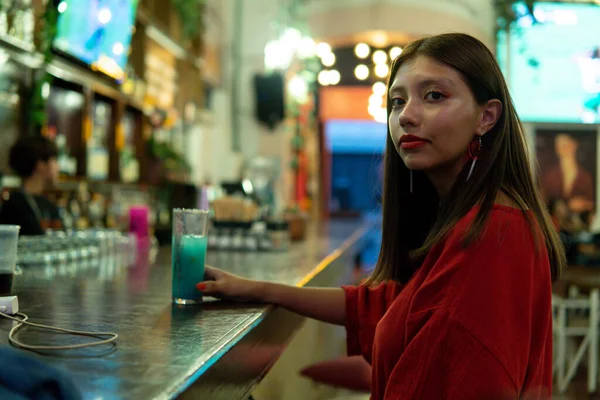 A young Latin woman enjoying a drink at a bar alone. Girl in a red dress with an alcoholic drink at the bar counter at night. There are backlights. She has a blue drink in her hand.