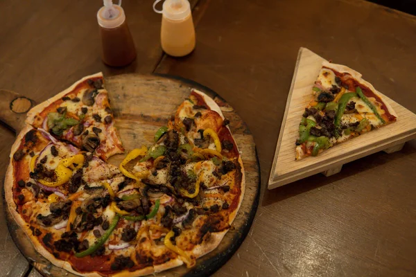 View from above of a wood oven pizza with mushrooms, meat, and green and yellow peppers. Piza with a slice cut and served on a wooden plate. Wood background.