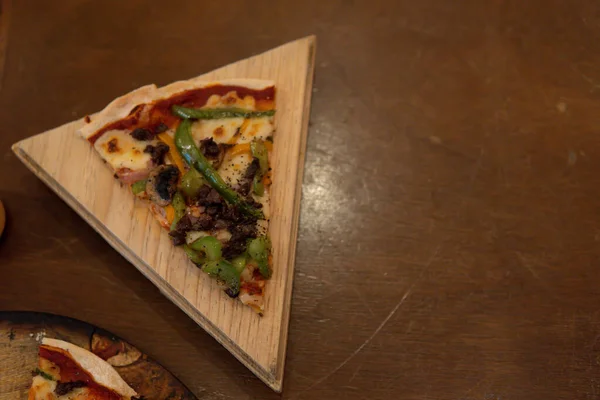 Piece of pizza with mushrooms and green pepper. Pizza slice on a wooden triangular plate and a wooden table. Freshly baked pizza is served on the plate.