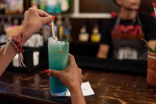 Hands of a girl mixing her alcoholic drink at a bar counter next to the bartender. Blue drink in the hands of a girl with bracelets. Counter of a bar with a waiter and a client.