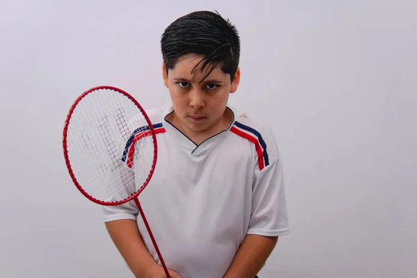 Boy in a sports shirt with a challenging look playing badminton. Athlete boy posing with a racket. Boy dressed in a sports shirt on an isolated white background with a racket in his hand.