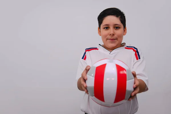 Boy dressed in a white sports shirt holding a volleyball ball on an isolated white background. Boy showing his white and red volleyball ball.