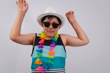 Plaj kıyafetleri içinde beyaz bir arka planda çiçek kolyesi takan mutlu bir çocuk. Sahilde kolları havada çok hareketli bir çocuk. Kısa kollu mavi gömlekli ve beyaz şapkalı çocuk..