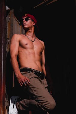 Athletic latin fitness model posing shirtless with hat and sunglasses in a rusty metal train car. Man with a confident attitude posing in an abandoned train.
