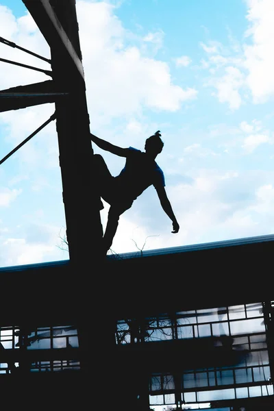 Black silhouette of a man climbing a metal structure. Black man doing an extreme activity. Silhouette of a man on top of a tower posing with the sky in the background.