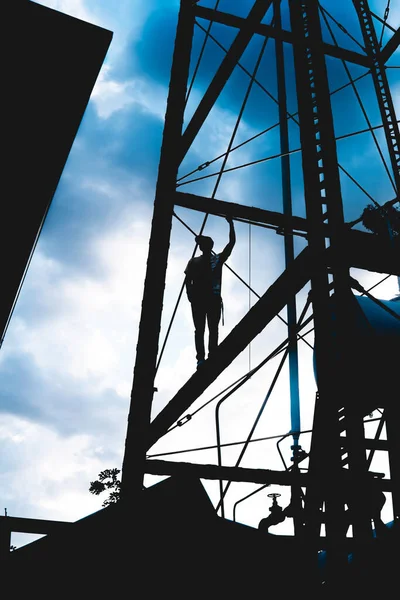 Boy on top of a metal tower posing with the sky in the background. Black silhouette of a man on a tall metal structure doing extreme activities. Shadow of a man climbing on tall buildings.