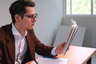 A businessman works in his white office. Elegant Caucasian man reading his black notebook. Business executive analyzing data from a graph. Businessman dressed in a suit, with glasses and shirt. High