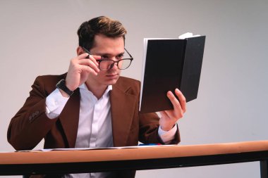 Man with glasses reading his black notebook. Man adjusting his reading glasses. Caucasian and blond man dressed in a suit and with glasses at a desk and with a white background. High quality photo