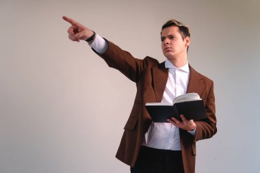 Teacher dressed in a suit on a white background holding a book while pointing somewhere. Business man pointing with a book in his hand. Lawyer pointing accusingly.