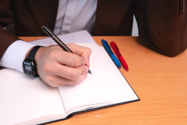 Desk where a man in a suit is writing. Wooden desk with pens and a notebook where a lawyer is writing. Man writing in a black notebook.