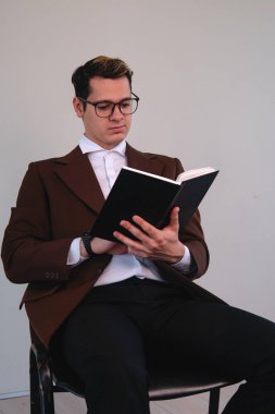 Studious elegant man sitting in front of a white wall while reading a book. Businessman enjoying a good read. High quality photo