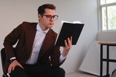 Elegantly dressed man in a suit reading a book sitting in a white room. Man with brown coat and glasses studying. Business man reading. University professor with sack in a white room. High quality