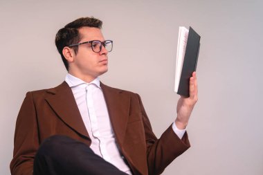A university professor on an isolated white background sitting attentively reading a book. An elegant man studying. Young man dressed in brown suit and wearing glasses. High quality photo