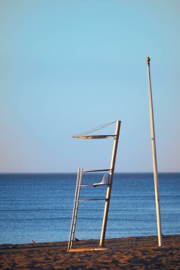 Sahilde gün doğumu, arka planda mavi deniz ve cankurtaran kulesi ayrıntıları var..