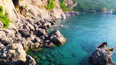 Şafak vakti kıyı şeridi, sahil kıyısındaki dağ ve kayalar, turkuaz deniz ve yeşil ağaçlar, devasa taş yapılar, tatil köyünün güzel hava manzarası.