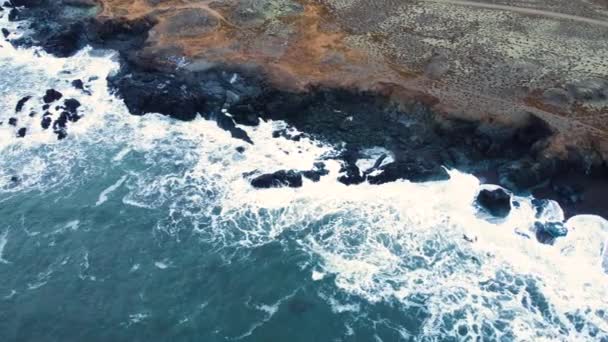 Aerial top view of waves break on rocks in a blue ocean. Sea waves on beautiful beach, Birds eye view of ocean waves crashing against an empty stone rock cliff from above, Iceland — Stock Video