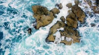 Deniz kayalık sahili yıkar, havadan çekilen karenin üstünde, sahil şeridinde uçar, Kayalıklarda Mavi Saf Suyla, Okyanusta güneşli havada. Kuş Gözü Görünümü 4K Sinematik İnsansız Hava Aracı. Havadan Gelen Sahil, 