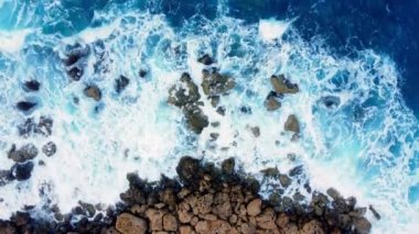 Deniz kayalık sahili yıkar, havadan çekilen karenin üstünde, sahil şeridinde uçar, Kayalıklarda Mavi Saf Suyla, Okyanusta güneşli havada. Kuş Gözü Görünümü 4K Sinematik İnsansız Hava Aracı. Havadan Gelen Sahil, 