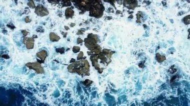 Deniz kayalık sahili yıkar, havadan çekilen karenin üstünde, sahil şeridinde uçar, Kayalıklarda Mavi Saf Suyla, Okyanusta güneşli havada. Kuş Gözü Görünümü 4K Sinematik İnsansız Hava Aracı. Havadan Gelen Sahil, 