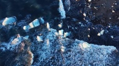 Saf mavi okyanus dalgaları ve ışık yansımaları İzlanda 'daki volkanik plajda buzdağlarını ezer. Yüksek kaliteli 4K görüntüler.