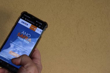 Taubate, SP, Brezilya - Temmuz 07 2022: ekranda CEF Caixa Economica logosu olan akıllı telefonu tutan eller. Sarı arka plan. Gölgeler.