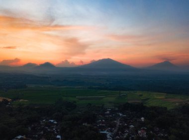 Pirinç tarlasının kırsal manzarası ve gün doğumunda arka planda dağlar. Gökyüzü bulutlu havada turuncudur. Orta Java, Endonezya
