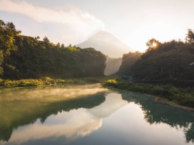 Su yüzeyi yeşilimsi görünen ve sabahları duman çıkaran bir göl ve gün doğumunda arka planda bir volkan. Göl ağaçlarla çevrili. Endonezya 'da Merapi Volkanı adında bir volkan..