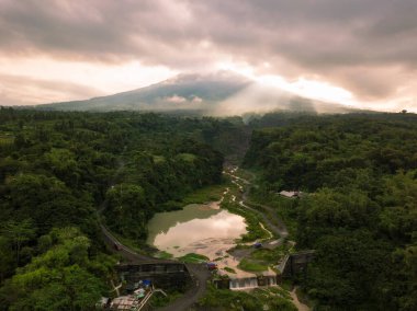 Bebeng Nehri ile Merapi Dağı manzarası ve su tutan bir göl, gökyüzü bulutlu görünüyor. Gölün etrafını saran ağaçlarla dolu orman bitkileri. Bego Pendem, Merapi Dağı