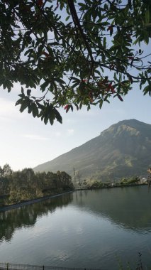 Yanında ağaçlar olan bir göl. Güneşin ışınları sabah yaprakların arasından açık bir gökyüzü ve az sayıda bulutla kırılarak görülebilir. Embung Kledung adında bir park. Endonezya 'da bulunmaktadır.