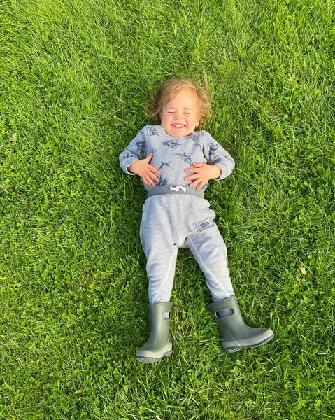 快乐的孩子蹒跚学步的男孩躺在外面的草地上 穿着橡胶靴 — 图库照片