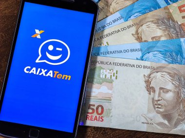 Smartphone with app (CAIXA TEM) on the screen with Brazilian real notes. Brazilian Government Emergency Aid on Caixa Bank App.