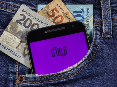 Ekranda, üzerinde 100 reais, 200 reais ve 50 reais olan kot pantolon cebinde Bank Nubank logosu olan akıllı telefon. Nubank bir finans şirketi..