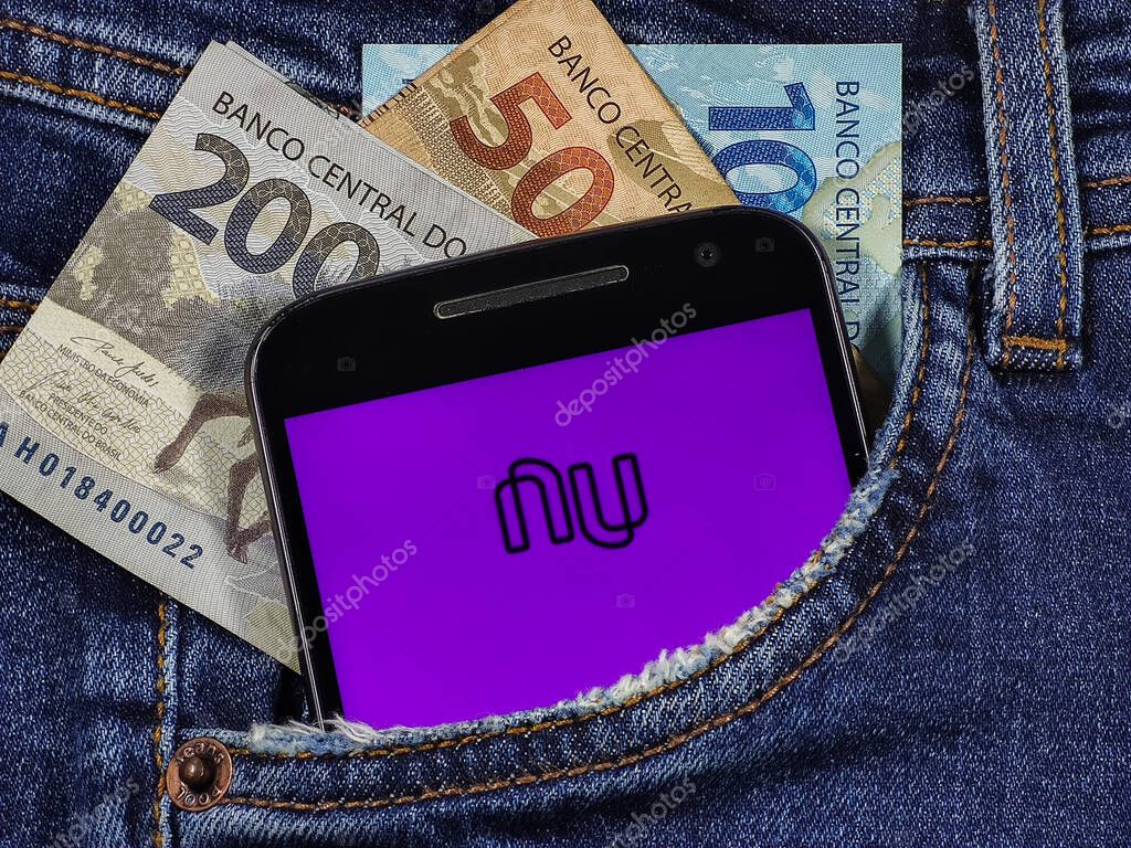Smartphone with Bank Nubank logo on the screen in jeans pocket with bills of 100 reais, 200 reais and 50 reais. Nubank is a financial services company.