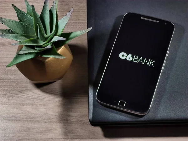 Akıllı telefon ekranındaki C6 Bank logosu ahşap masadaki defterin üstünde. C6 Bank Brezilya 'nın dijital bankasıdır..