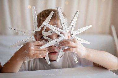 Beyaz tişörtlü küçük çocuk evde beyaz boyalı çamaşır mandallarından tahta kar taneleri boyuyor, elleri boyalı, geri dönüşümlü süs, yeni yıl dekoru, tek renkli fotoğrafçılık, el sanatlarına odaklan.