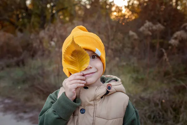 Niño divertido en sombrero amarillo que cubre un ojo con hoja amarilla durante el paseo de otoño de la familia — Foto de Stock