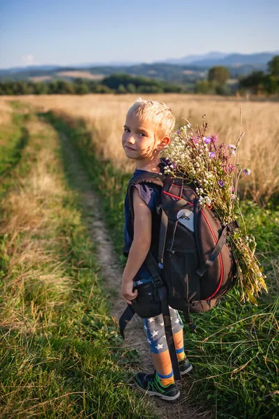 Маленький турист с рюкзаком приходит с горы и несет букет полевых цветов, красоты природы — стоковое фото