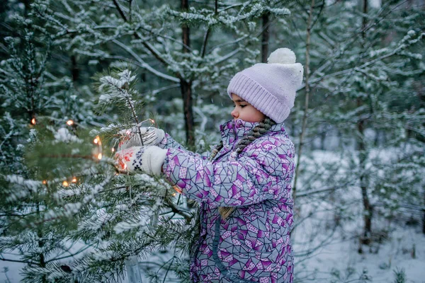 Дівчина, посміхаючись і сміючись, грає з сяючою гірляндою під час прогулянки в лісі на сніговий день — стокове фото