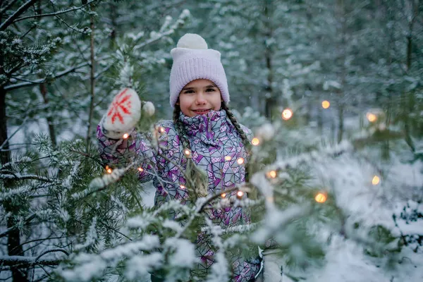 Dziewczyna, uśmiechnięta i śmiejąca się, bawi się świecącą girlandą podczas spaceru po lesie w śnieżny dzień — Zdjęcie stockowe