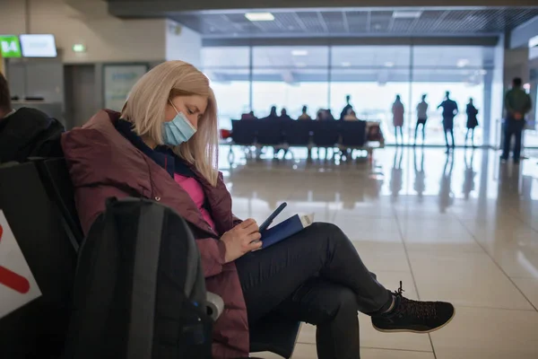 Solo traveler in medical mask sits and uses mobile in terminal while waiting for flight in airport — стоковое фото