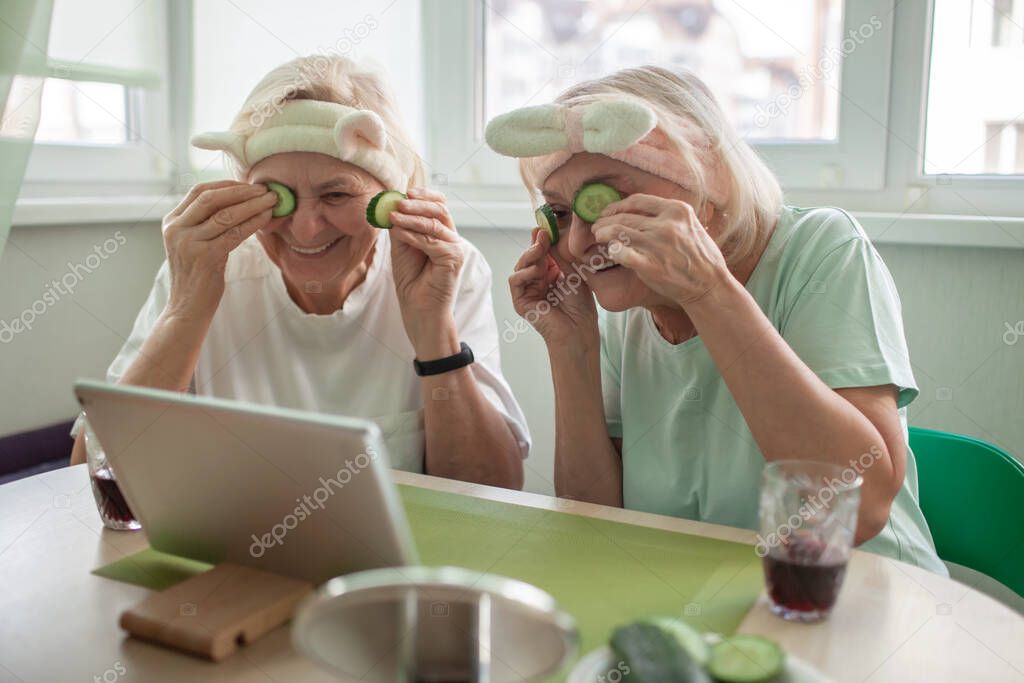 Two happy senior women apply facial cosmetic mask, have fun at home, body positivity, ageism
